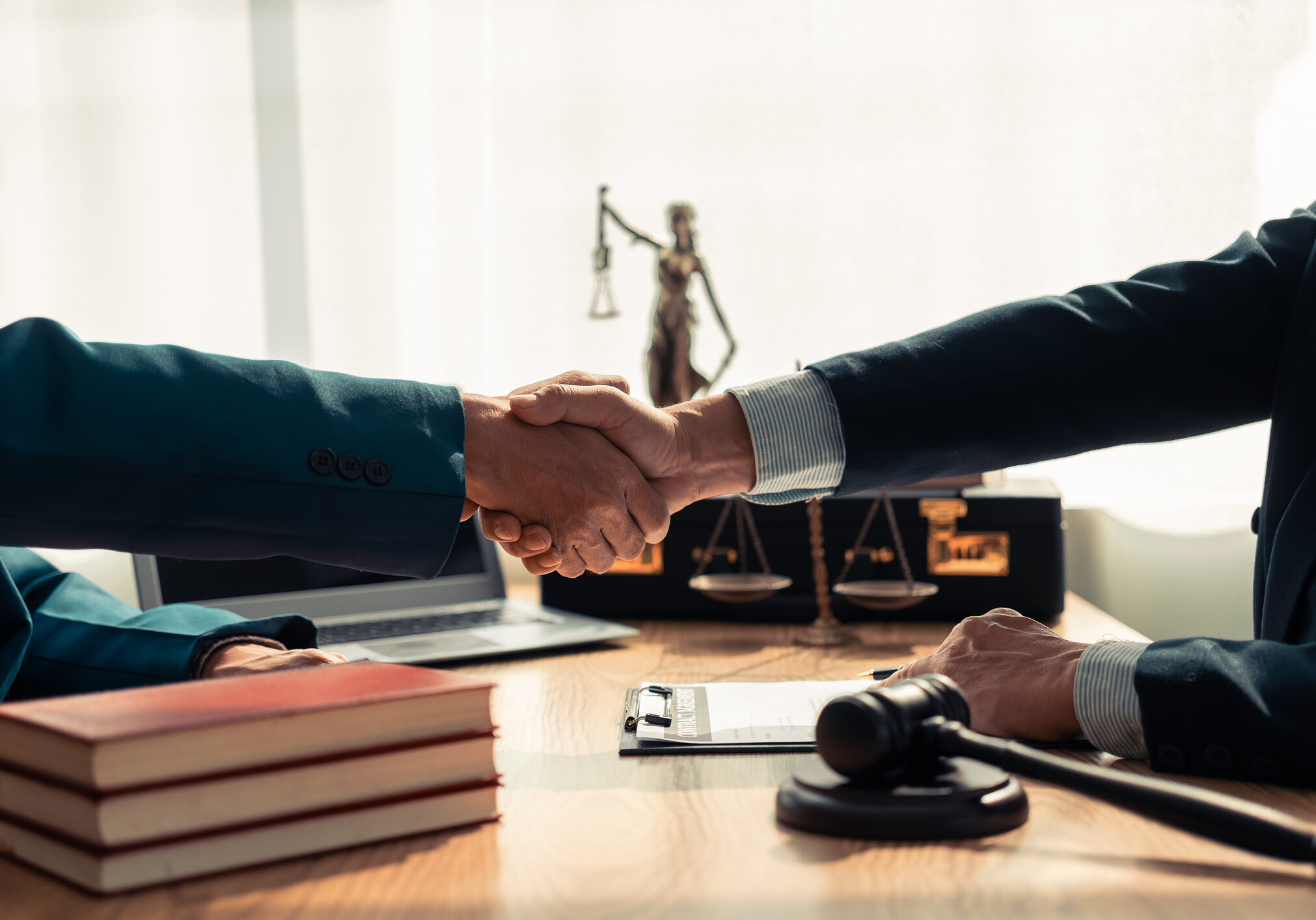 Close-up of lawyer and client shaking hands on table, symbolizing trust in legal contract with a hammer and a statue on the table, symbolizing justice.